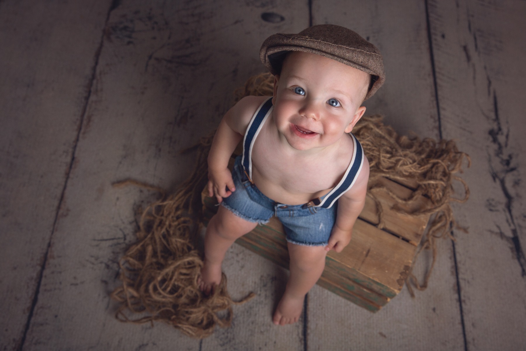 baby in a driver hat and suspenders sits on a wooden crate