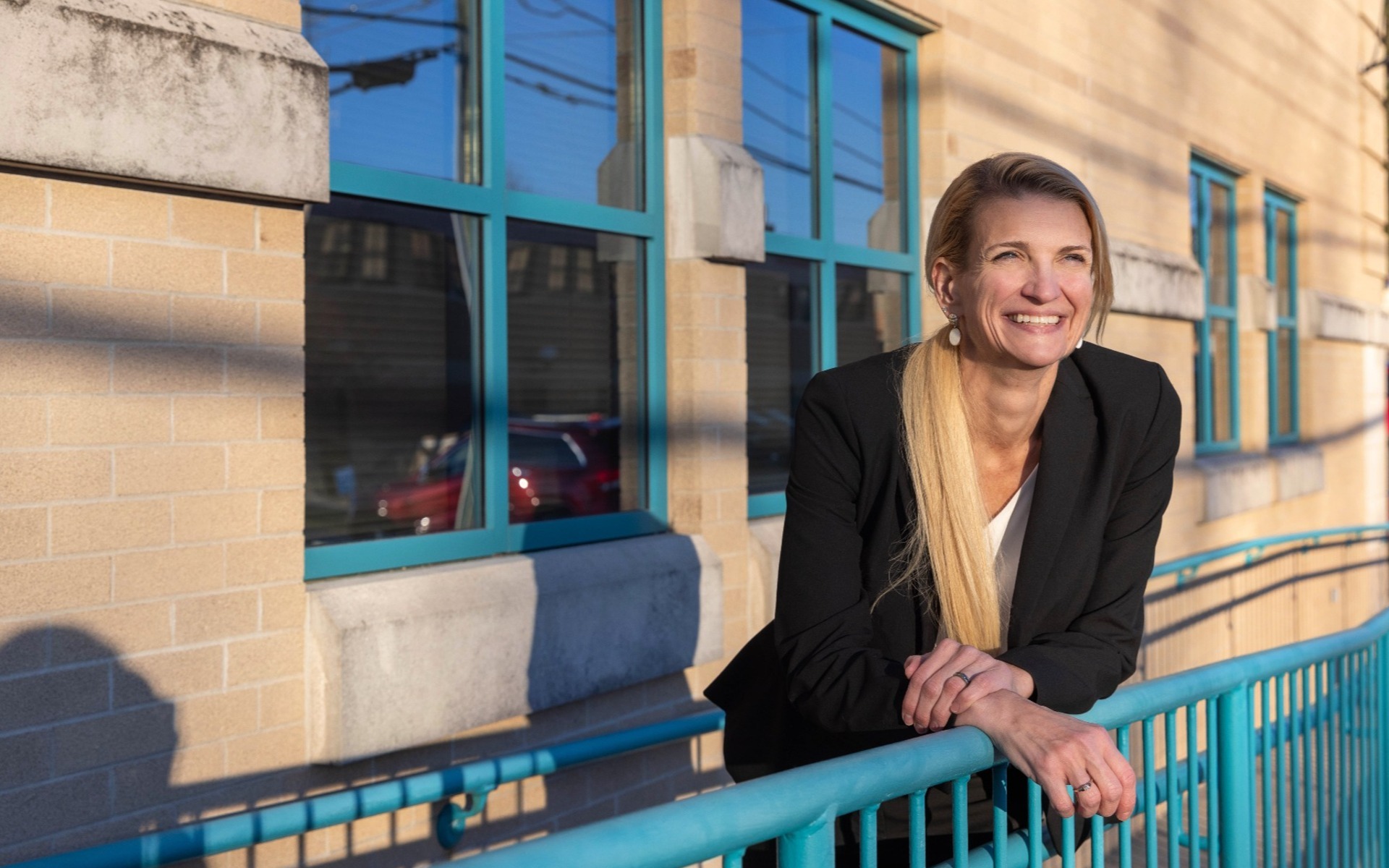 blond haired business woman looks off camera as she poses on a teal blue railing