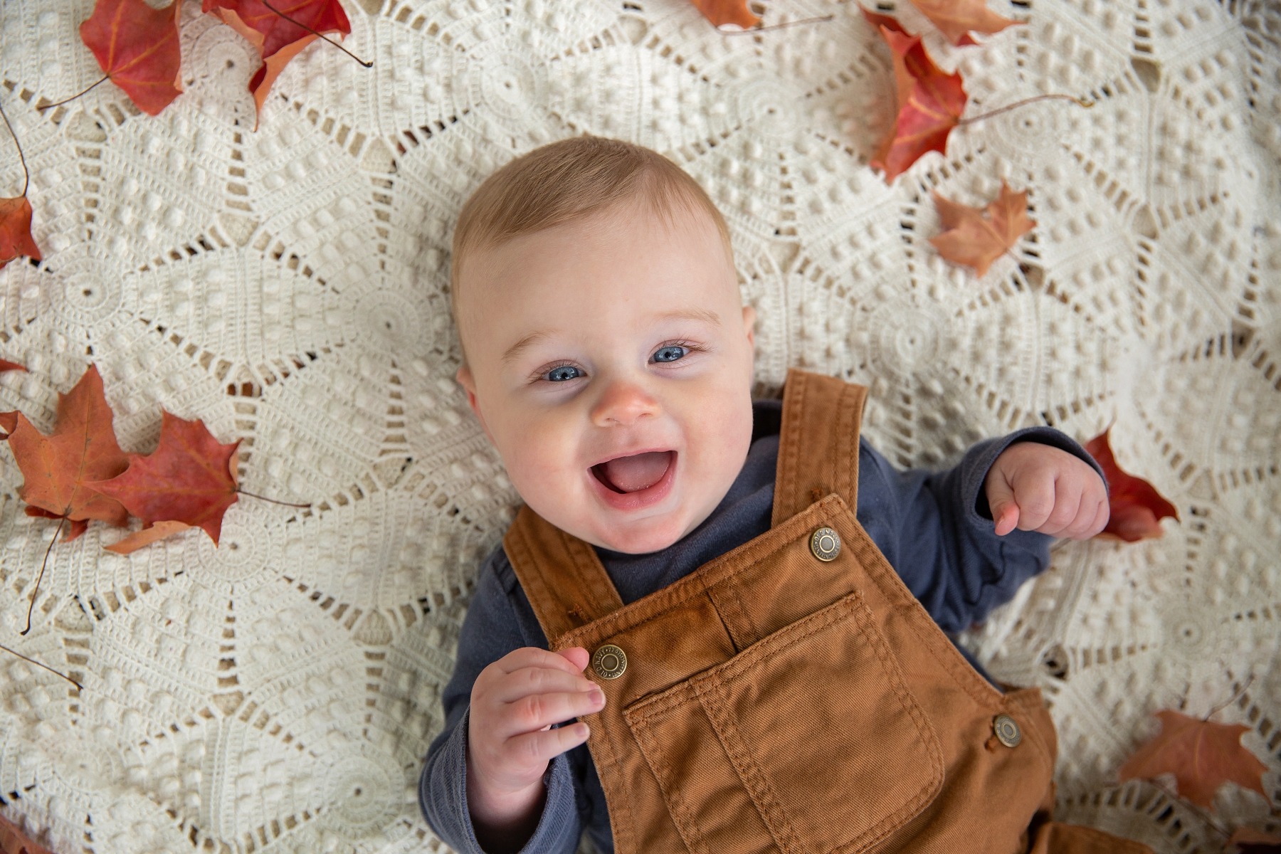 baby boy in carhart suspenders smiles up from a white blanket