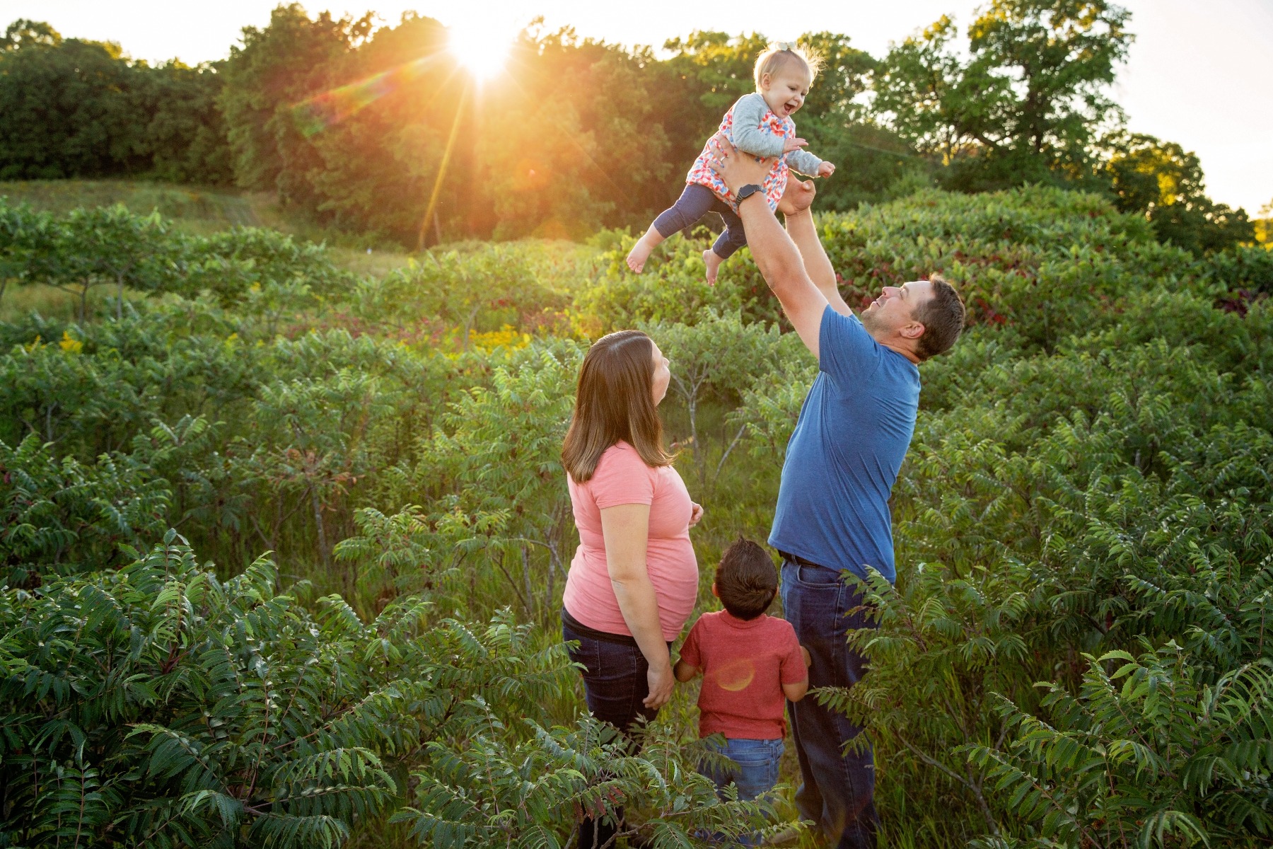 family of 4 on a green hillside, dad tosses baby girl in the air